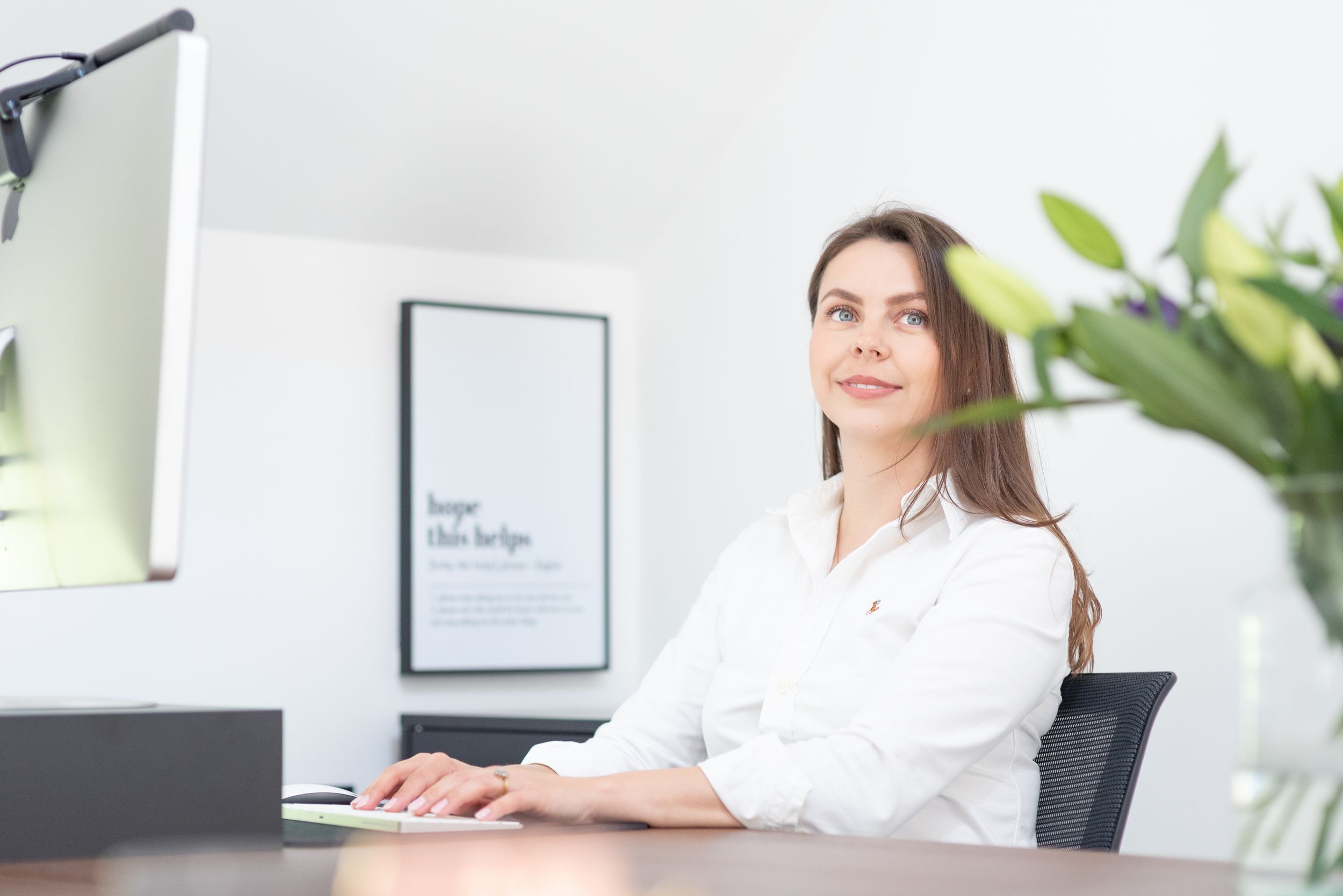 Professional in white blouse sits at modern office desk with houseplant and wall art in background.