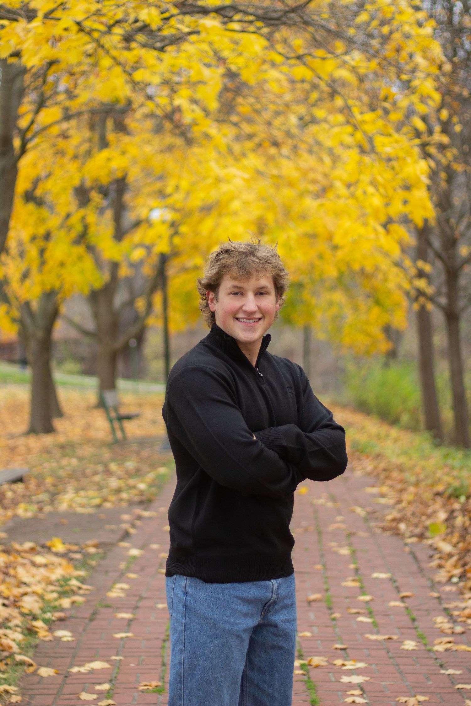 A person in a black sweater stands smiling on a brick path with vibrant yellow autumn foliage overhead during senior photos.