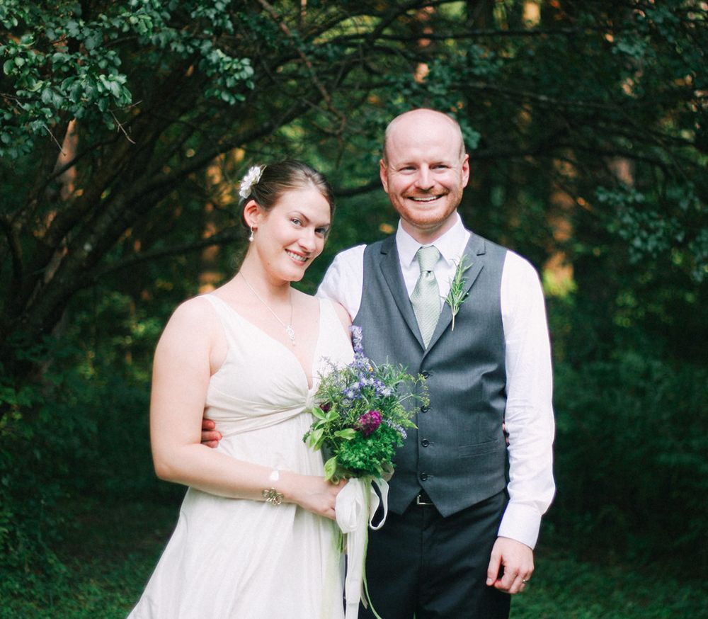 Smiling bride holding wildflower bouquet standing with groom in gray vest in a forest.