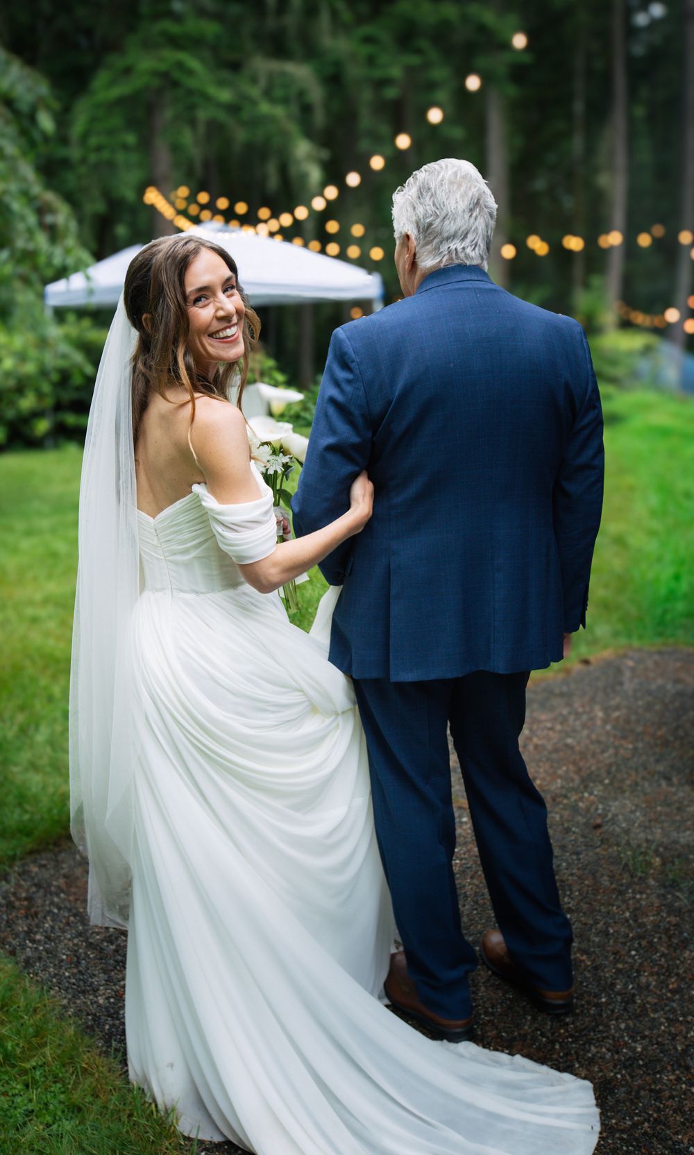 Smiling bride in white off-shoulder gown and veil walks with man in navy suit outdoors with string lights.