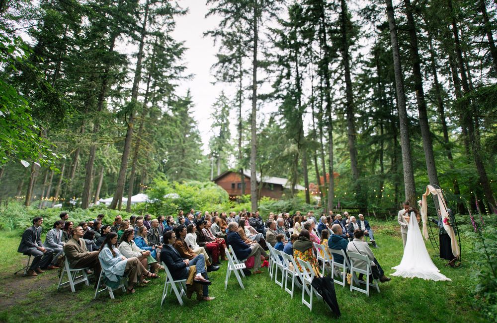 Outdoor wedding ceremony in a lush forest clearing with guests seated in white chairs facing the couple.