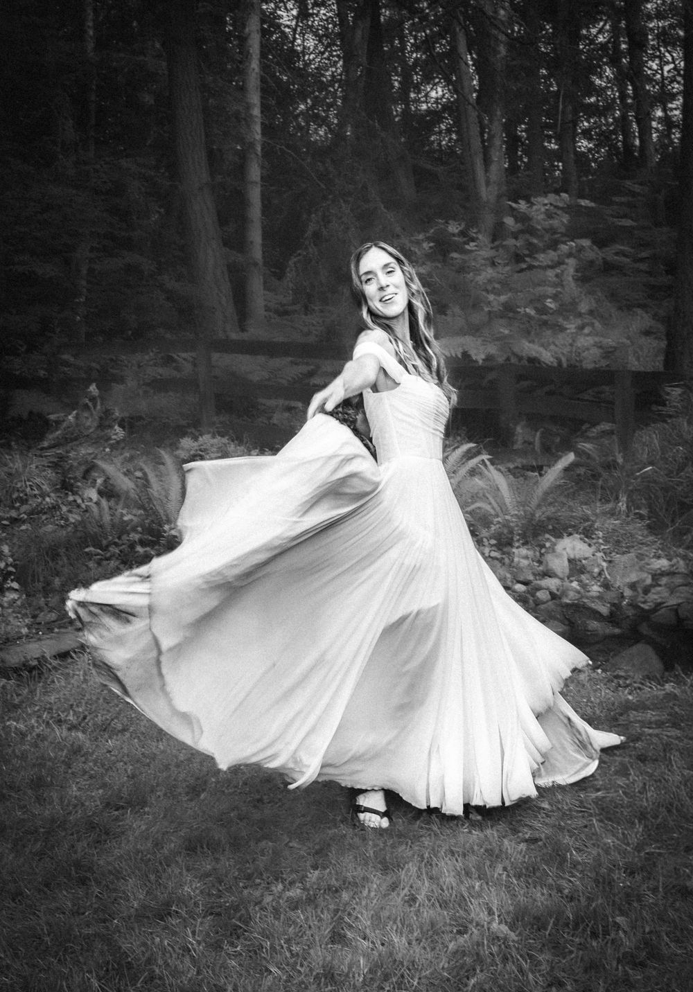 Black and white photo of a woman in a flowing white wedding gown twirling outdoors in a lush forest setting.
