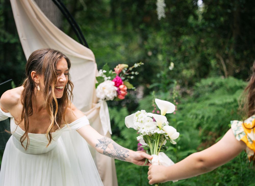 Smiling bride in white off-shoulder gown receiving a white floral bouquet outdoors at a garden wedding ceremony.