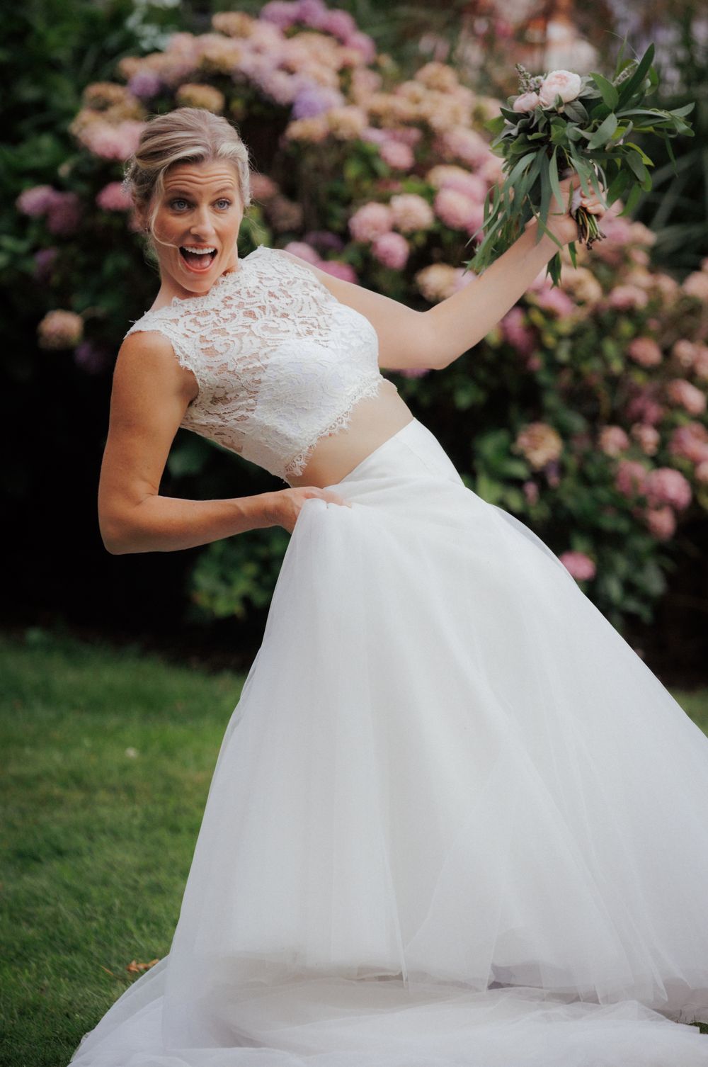 Joyful bride in two-piece lace wedding gown playfully posing in front of hydrangea bushes.