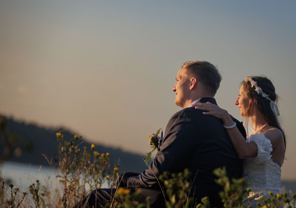 Bride and groom embracing while gazing at scenic waterfront view during romantic sunset wedding portrait.