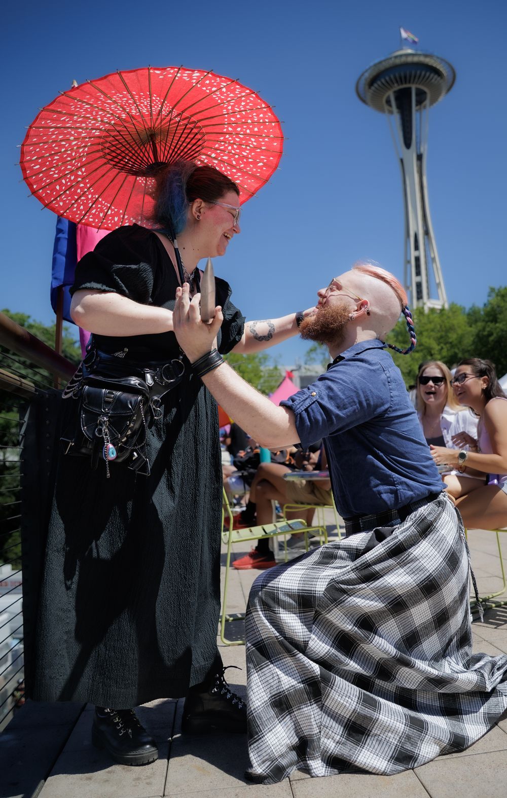 Man kneeling in plaid kilt proposing to woman with red parasol near Seattle Space Needle.