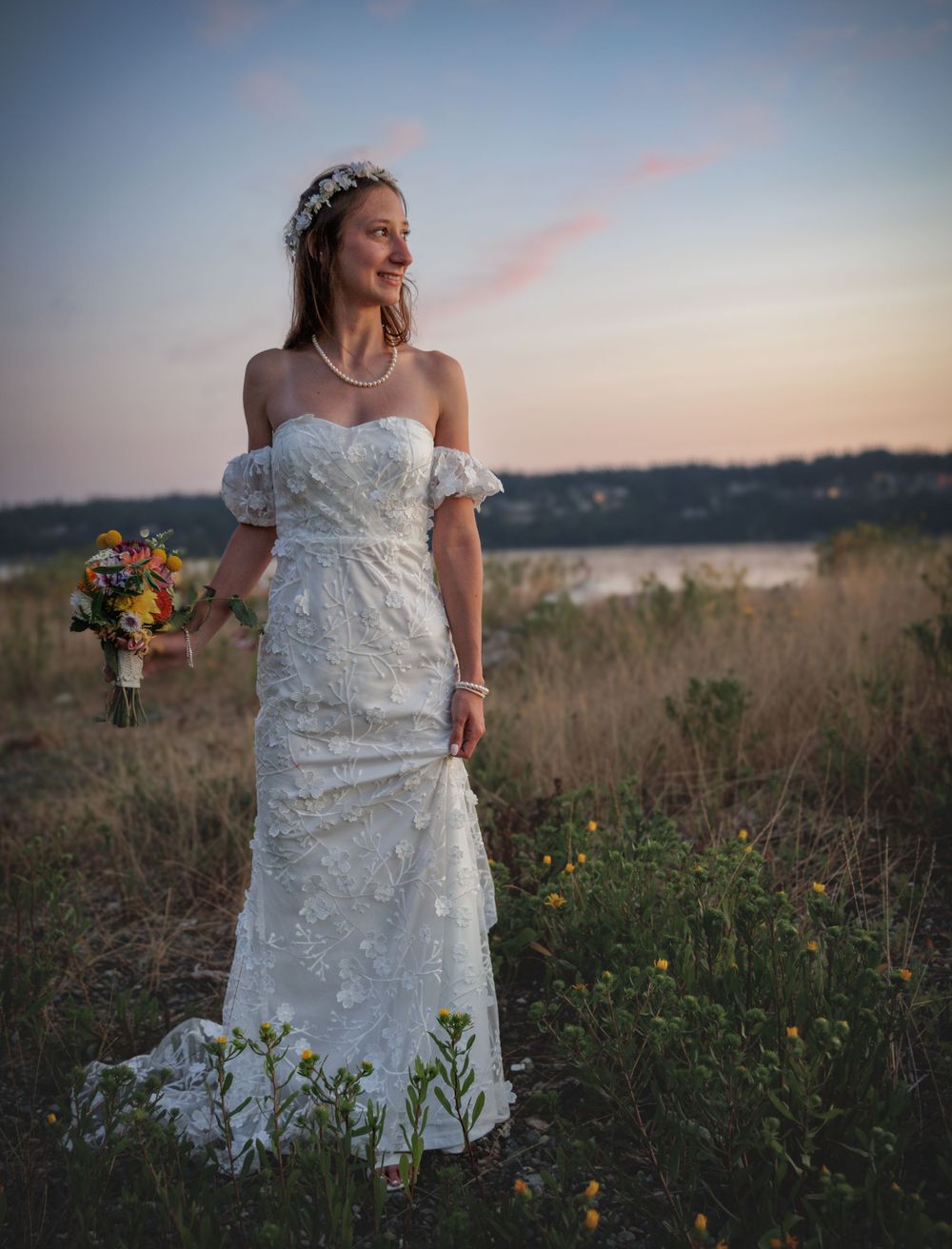 Bride in off-shoulder lace gown holding colorful bouquet posing in field during golden hour sunset.
