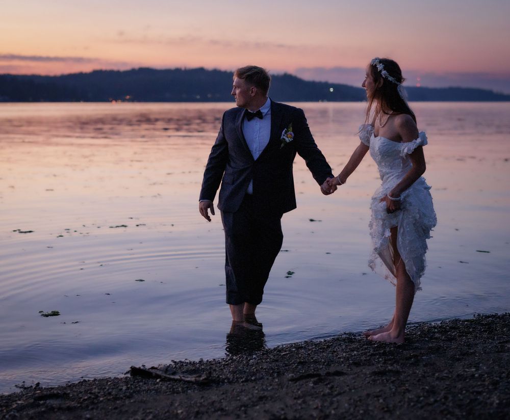 Bride and groom holding hands walking in shallow water at sunset by the ocean.