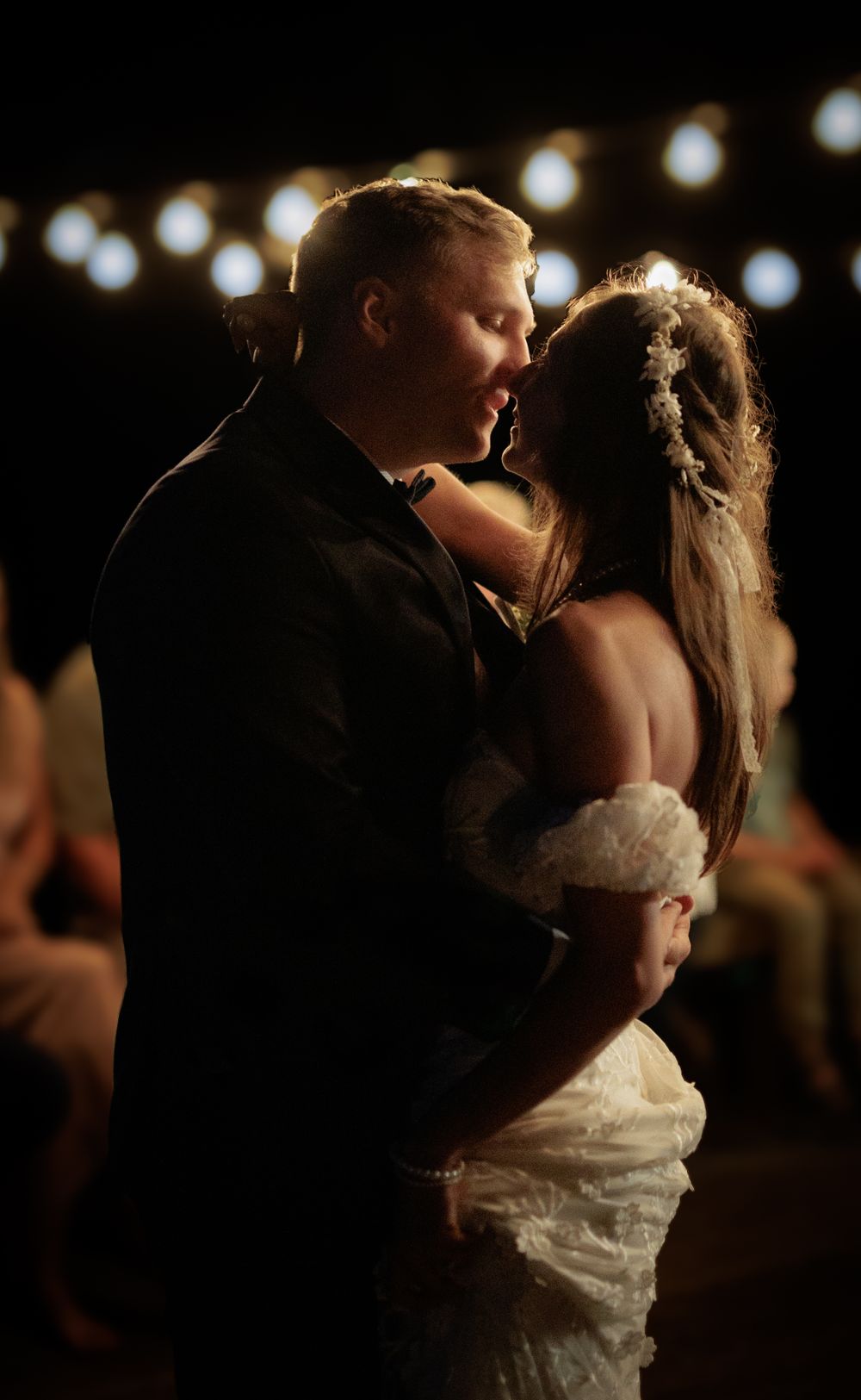 Bride and groom sharing romantic first dance under warm string lights during evening wedding reception.