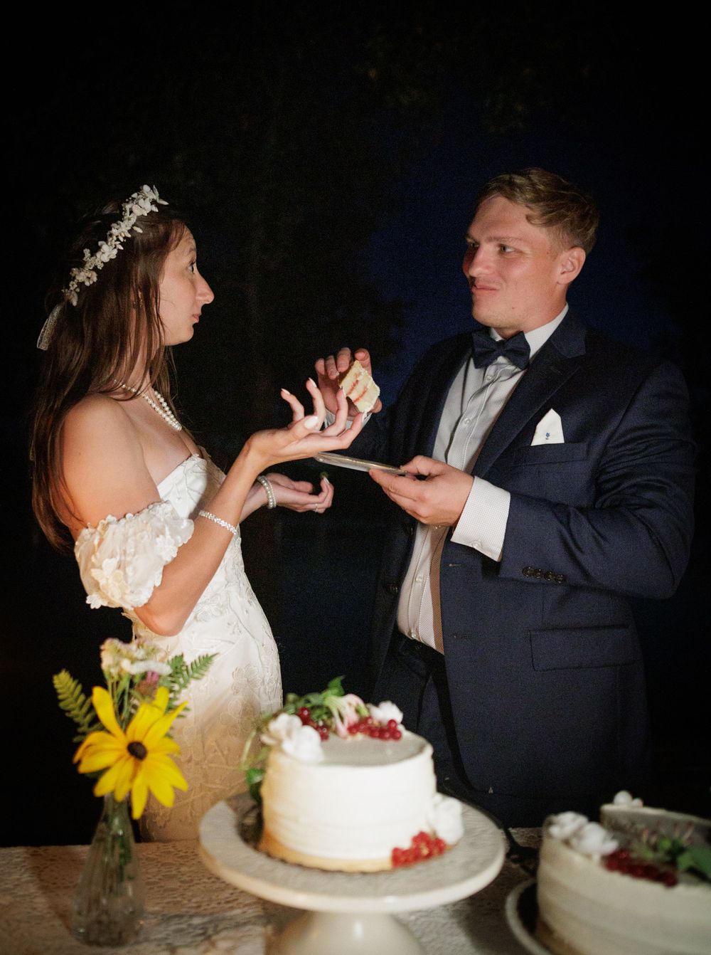 Bride and groom cutting white wedding cake decorated with berries during outdoor evening reception.