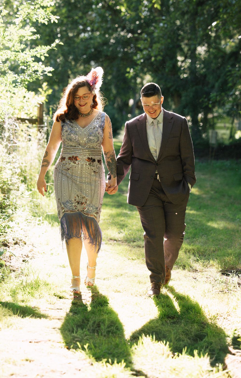A couple in formal attire walks hand-in-hand along a sunlit garden path, smiling joyfully.
