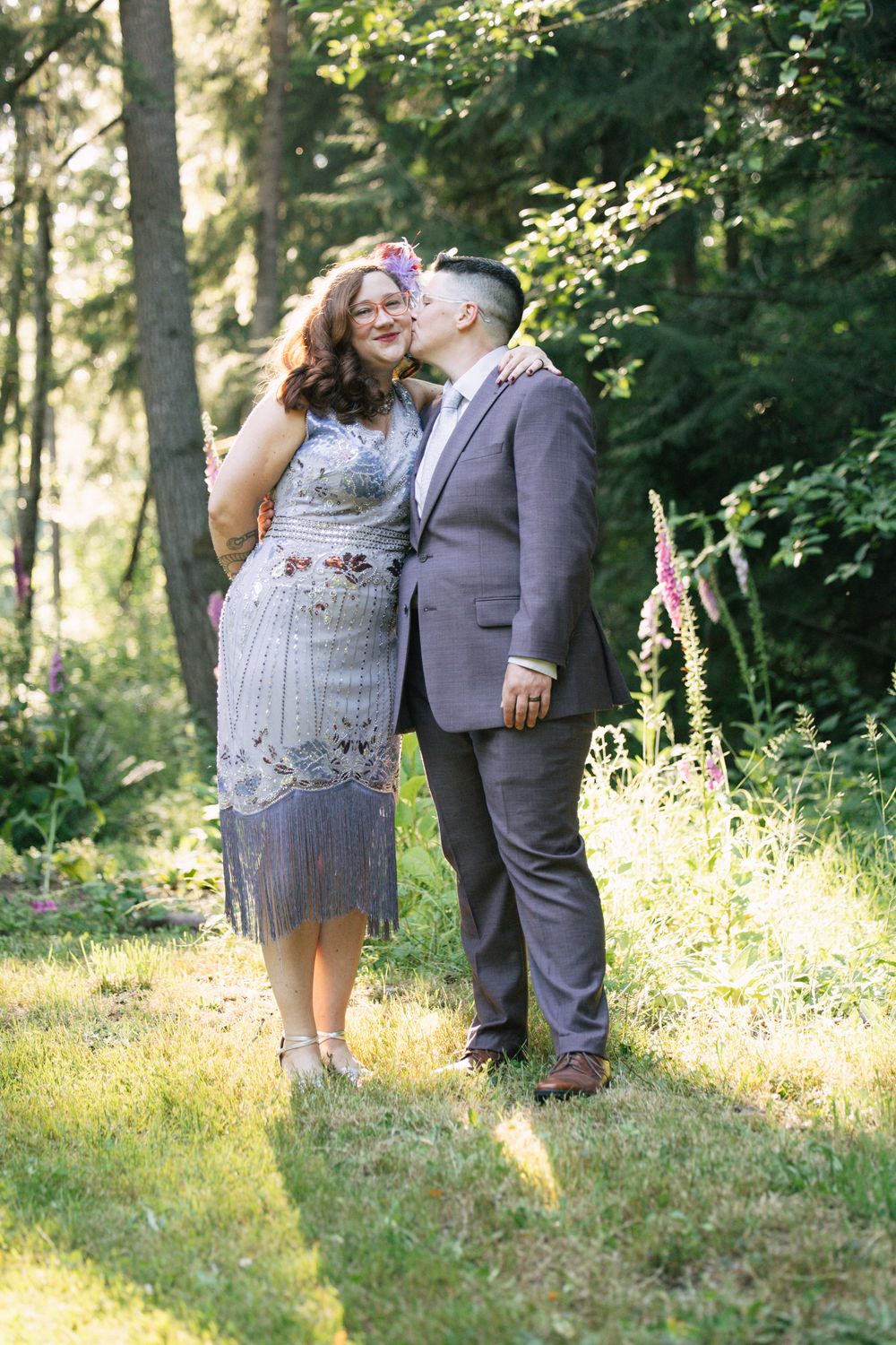 Couple sharing a romantic kiss in a sunlit forest garden, woman in embroidered dress, man in gray suit.