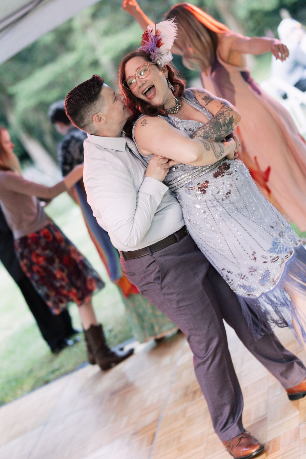 Couple dancing joyfully at an outdoor wedding reception on a wooden dance floor with guests in the background.