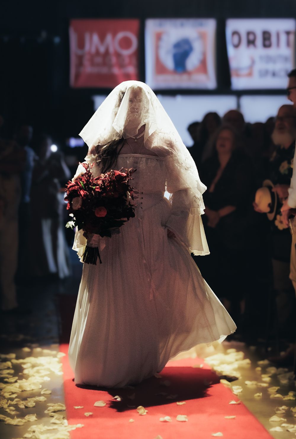 Bride in white gown and veil walking down runway holding dramatic dark red floral bouquet.