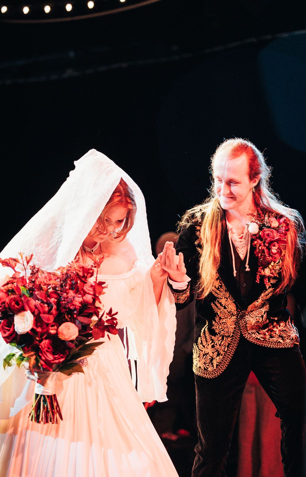 Bride in white veil holding red bouquet walking with long-haired partner in embroidered black jacket.