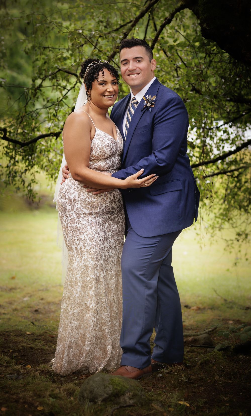 A smiling bride in lace gown and groom in blue suit pose together outdoors under green trees on their wedding day.