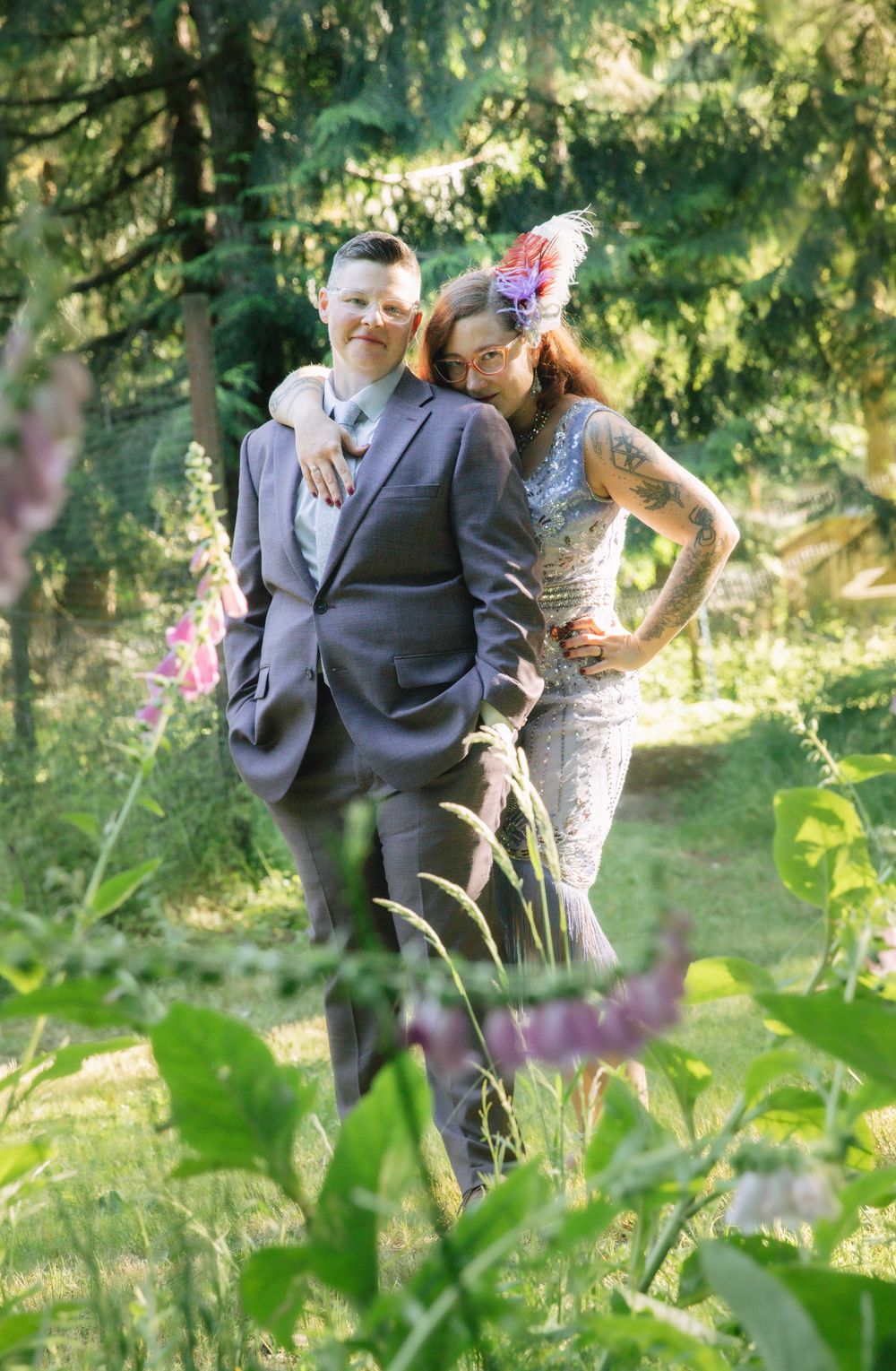 Couple in formal attire posing in a lush green forest, woman with tattoos and feathered headpiece kissing mans cheek.