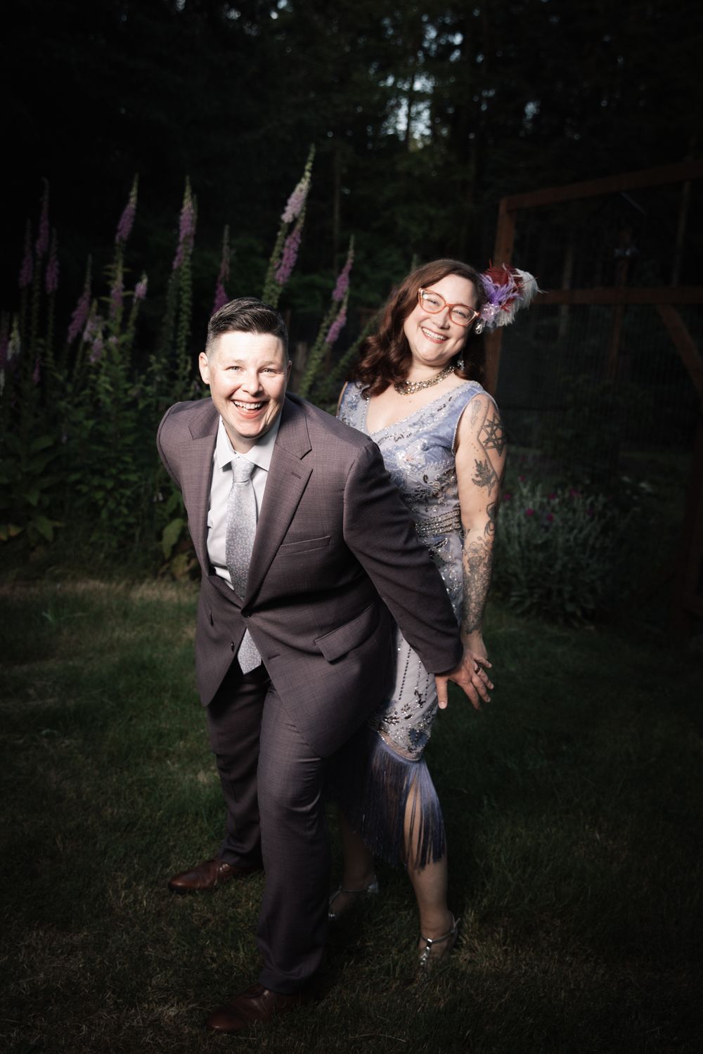 Two people in formal attire laugh joyfully outdoors, one in a gray suit and one in a blue lace dress with tattoos.