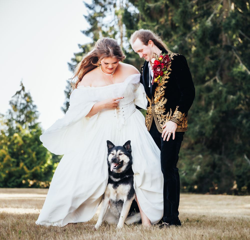 Wedding couple in elegant attire posing outdoors with their husky dog on a grassy field.