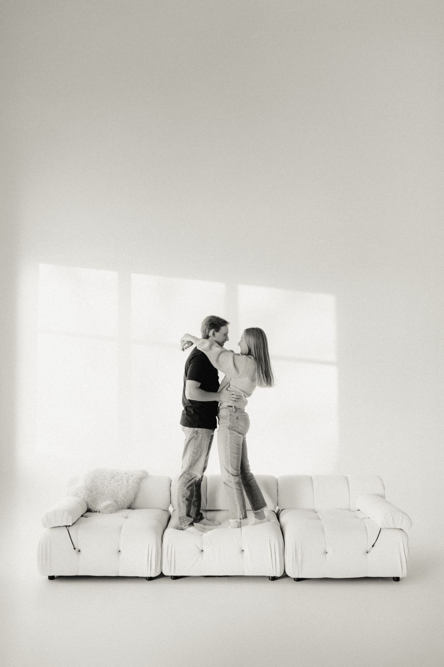 Black and white studio couples photo with a young couple on a couch, soft window light and timeless romantic posing.