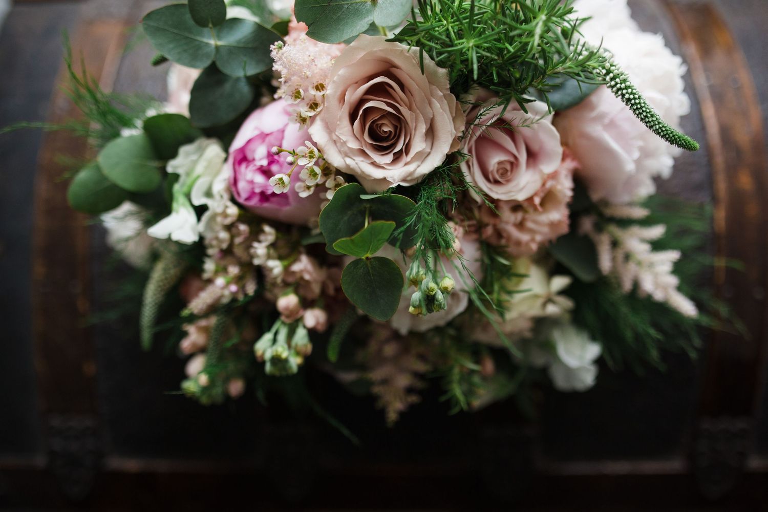 Elegant bridal bouquet with dusty pink roses, peonies, and lush greenery arranged on a rustic wooden surface.