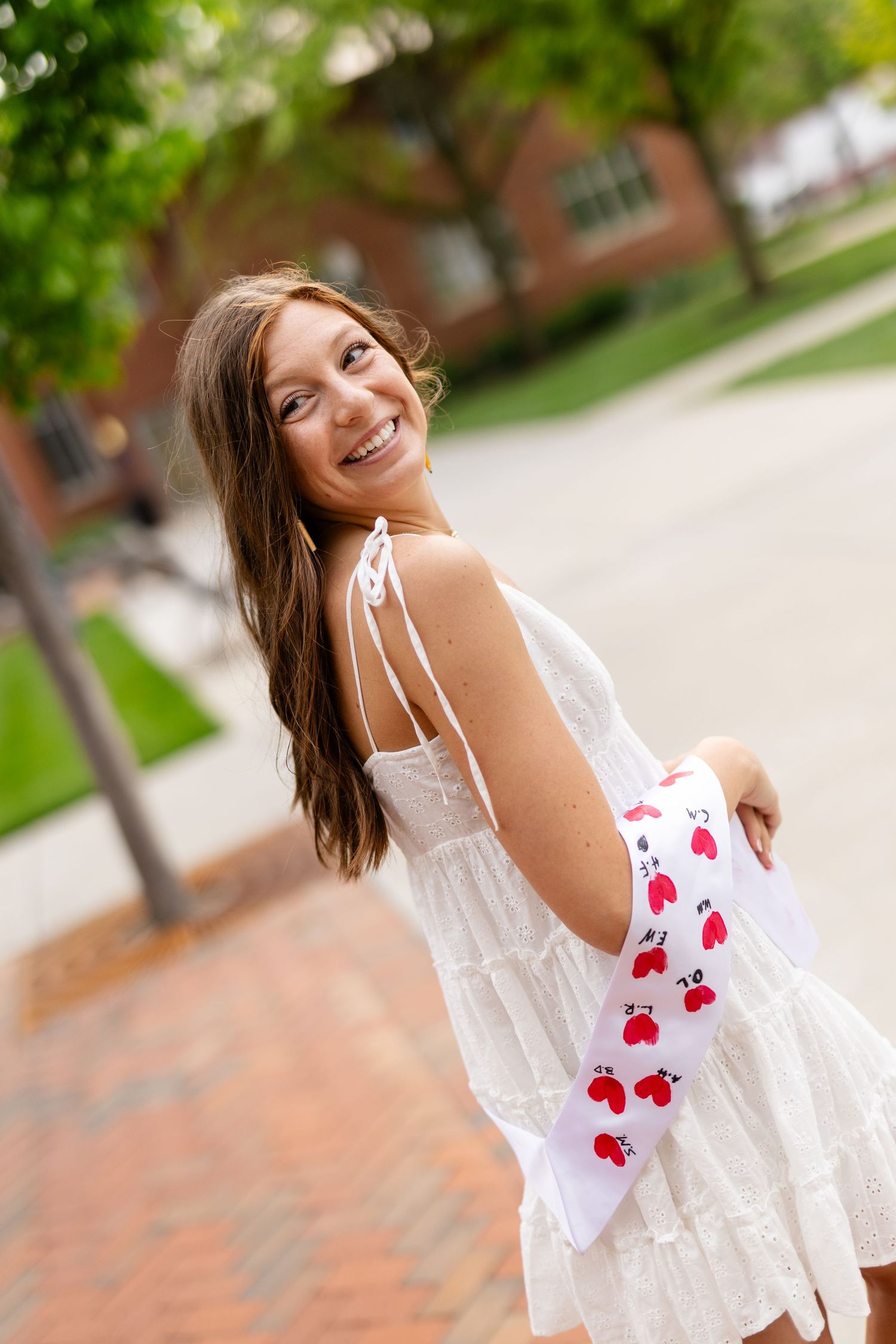 Smiling graduate in white dress wearing red heart sash and gold honor cord at outdoor graduation ceremony.