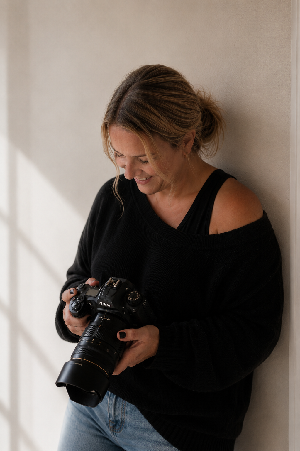 A female photographer smiles while reviewing images on her Nikon camera against a neutral wall.
