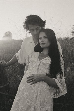 Black and white photo of a pregnant woman in lace dress embraced by a man in a field at dusk.