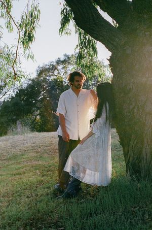 Couple standing by a large tree in warm sunlight, woman in white lace dress.