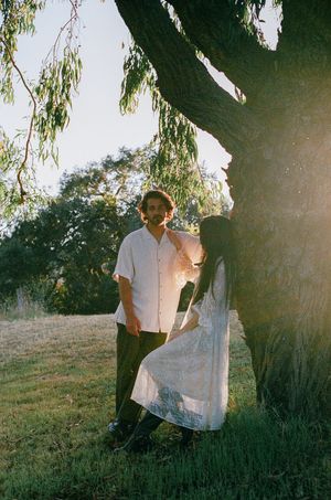 Couple posing under a large tree in a sunlit park, woman in white lace dress.