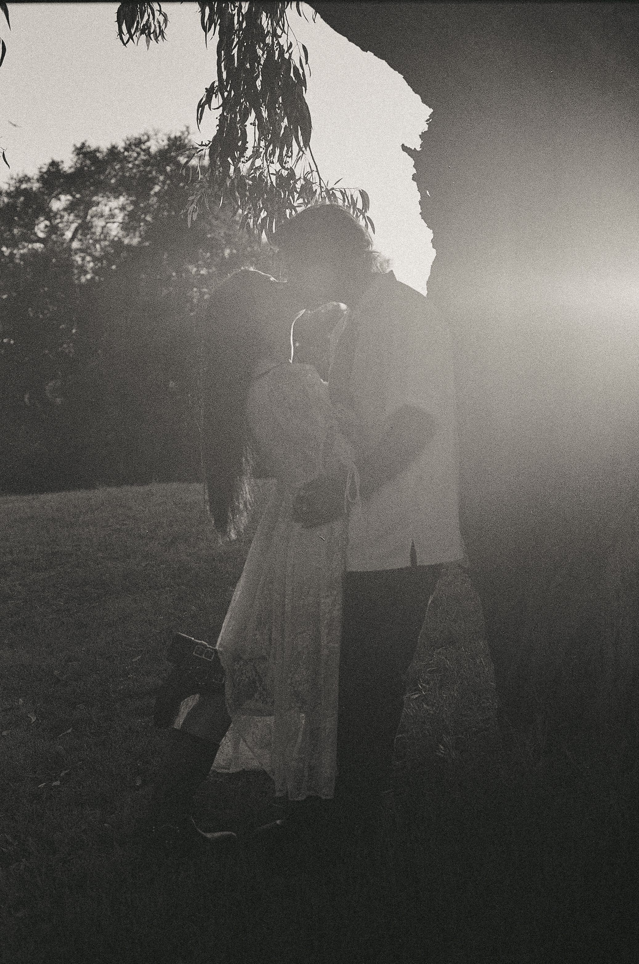 Silhouetted couple embracing in sunlit black and white outdoor portrait near rustic stone wall.