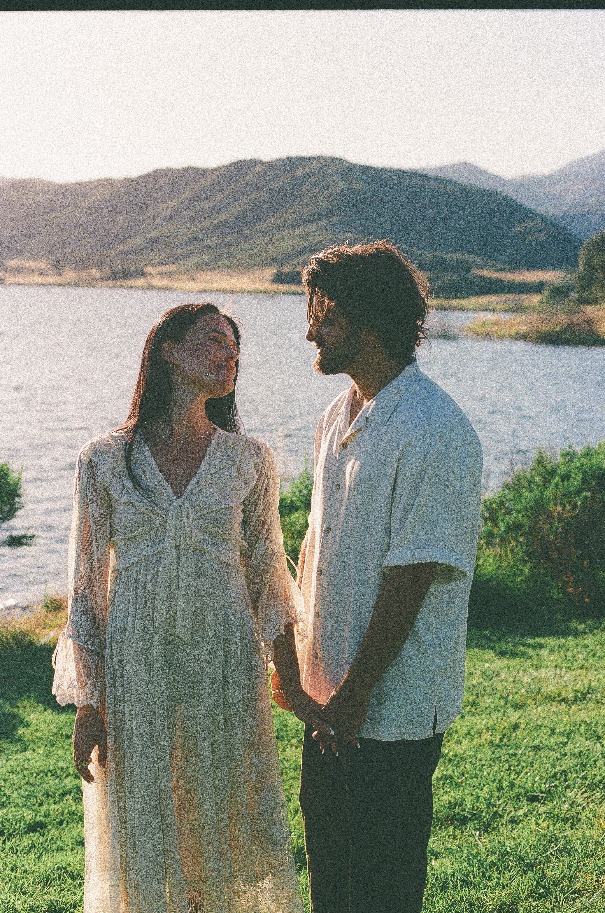 Couple in white lace dress and casual shirt standing by a lake with mountain backdrop in golden hour light.