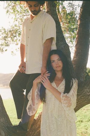 Couple in white outfits pose by a tree outdoors, woman in lace dress holds mans hand at golden hour.
