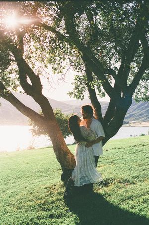 Couple embracing under a sunlit tree by a lake at golden hour, surrounded by lush green grass and mountains.