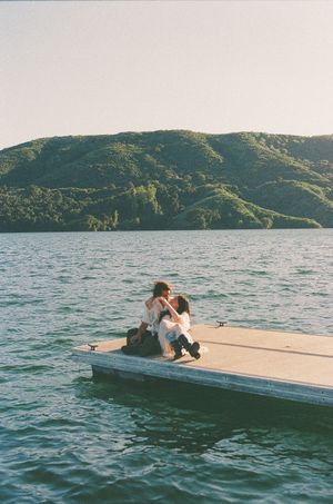 Two people embracing on a floating dock over a calm lake with lush green hills in the background.