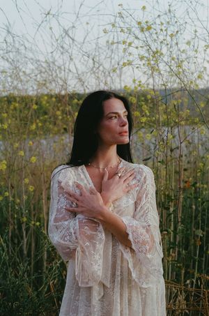 Woman in white lace dress stands in wildflower field, hands crossed over chest, gazing upward peacefully.