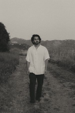 Young man in white linen shirt walking on a rural dirt path with misty hills behind him.