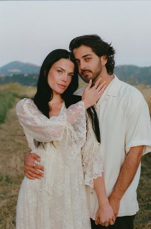 Romantic couple embracing in a field at dusk, woman in white lace dress and man in cream shirt.