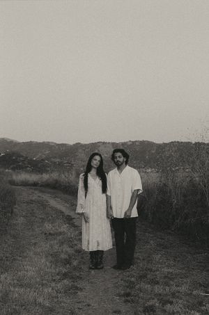 A couple in white clothing stands on a rural path surrounded by hills in a moody black and white photo.