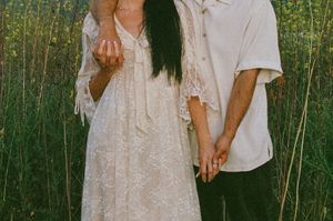 Couple holding hands in a field, woman in white lace dress, man in cream shirt, surrounded by tall green grass.