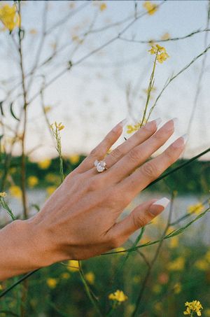 Close-up of an engagement ring on a hand among yellow wildflowers outdoors.