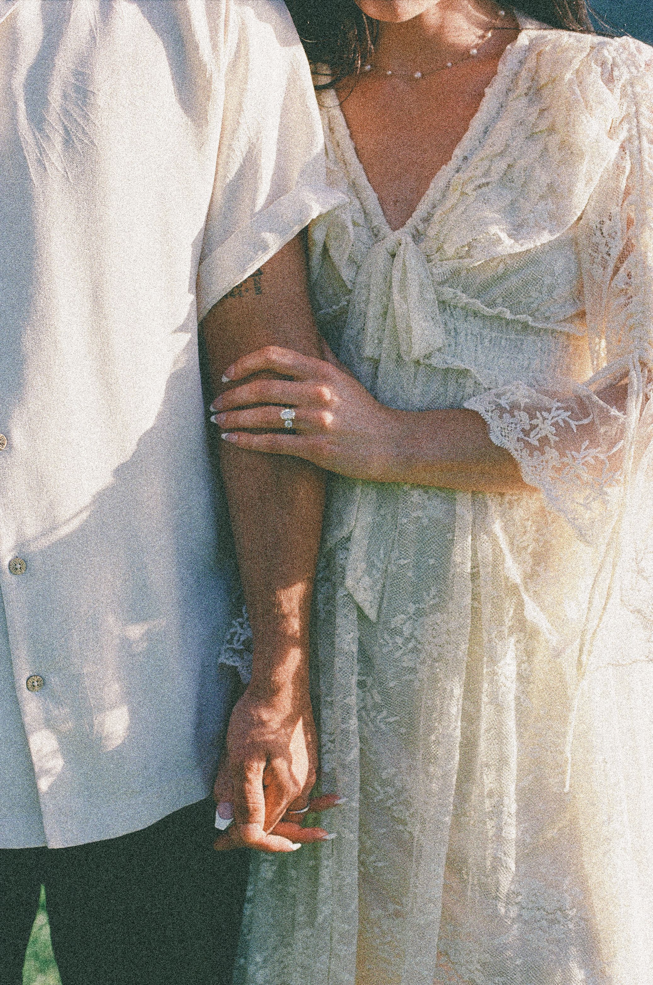 Couple in elegant white lace wedding attire holding hands, showcasing a diamond engagement ring in warm golden light.
