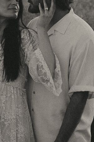 Black and white photo of a couple in elegant attire, woman in lace dress touching man's face, wearing an engagement ring.