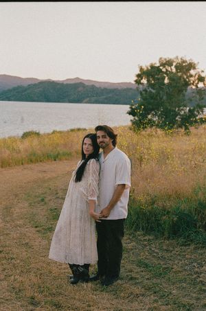 Couple posing together at golden hour near a lake with rolling hills, woman in white lace dress, man in white shirt.