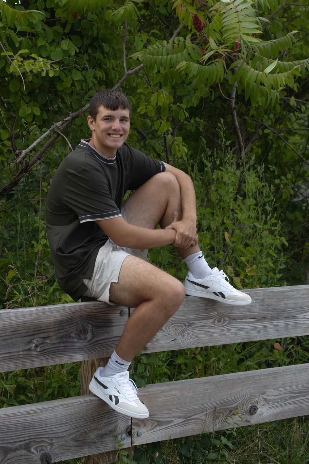 Smiling teenage boy in dark shirt and shorts sitting on a wooden fence outdoors.