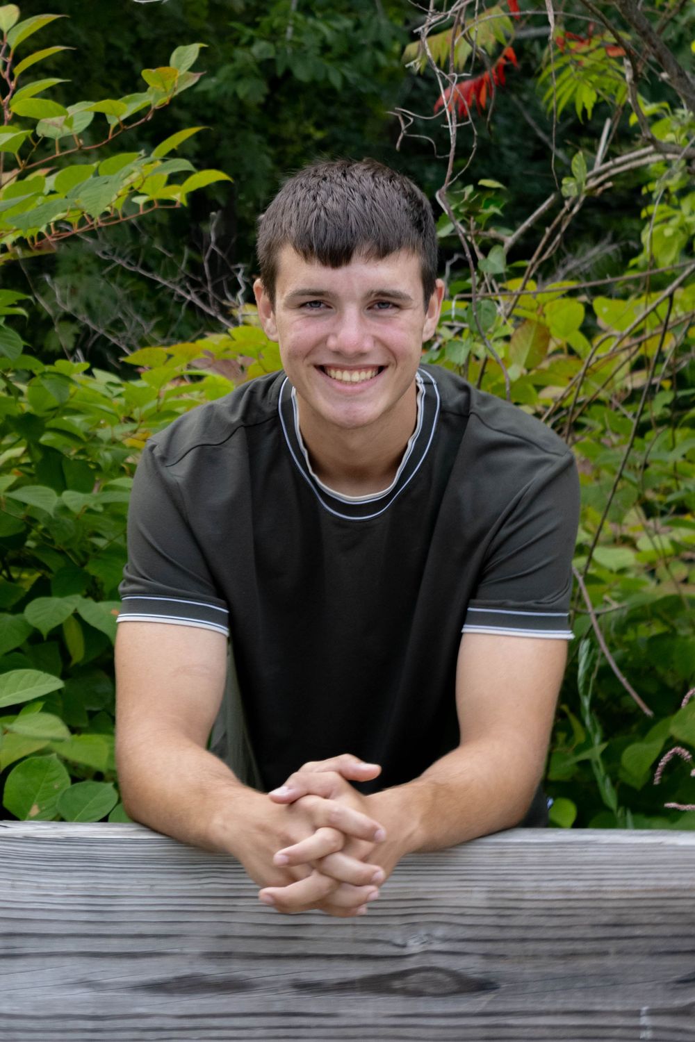 Smiling young man leaning on a wooden fence surrounded by lush green foliage.