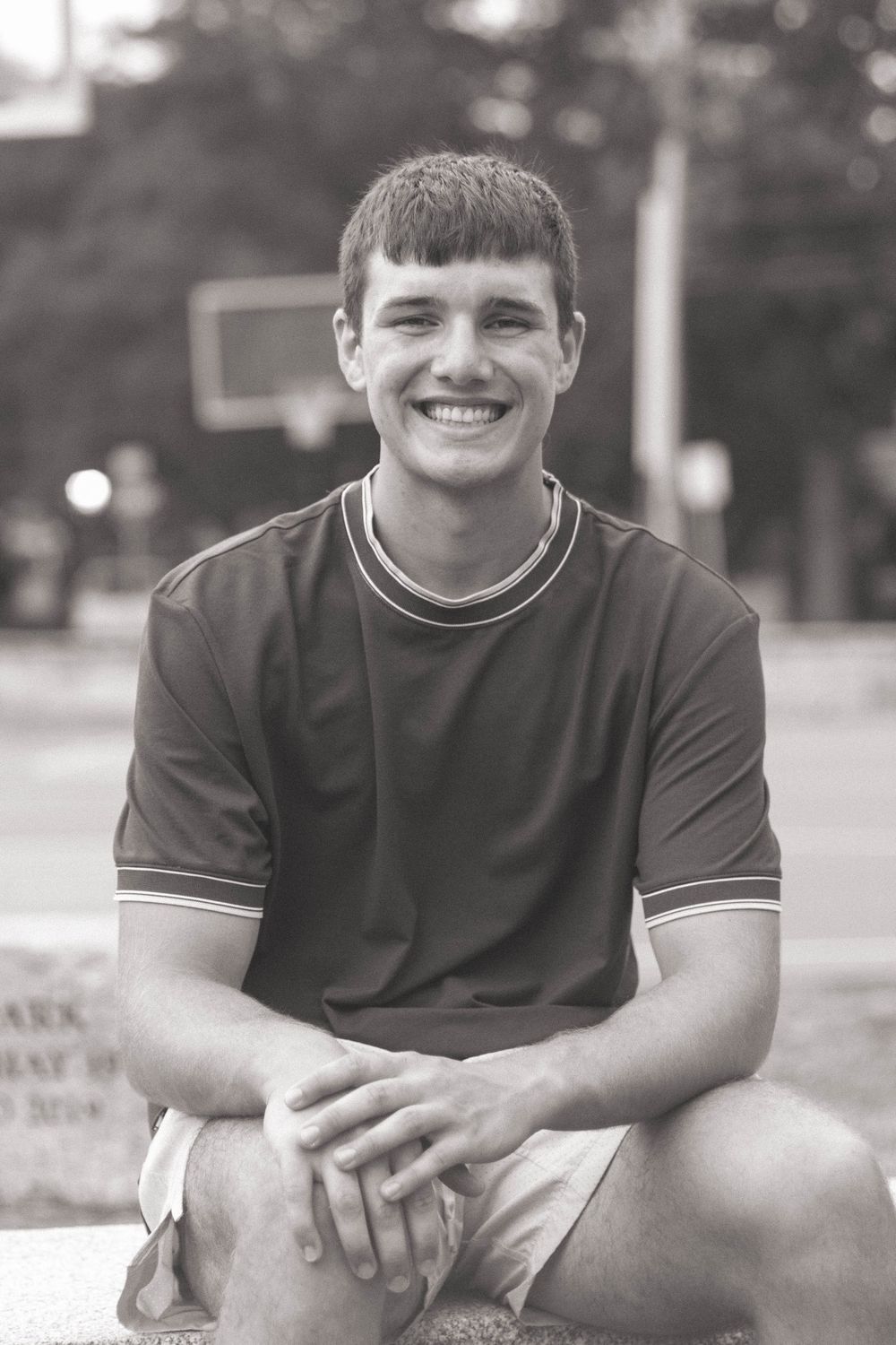 Smiling teenage boy in dark polo shirt sitting outdoors in black and white photo.