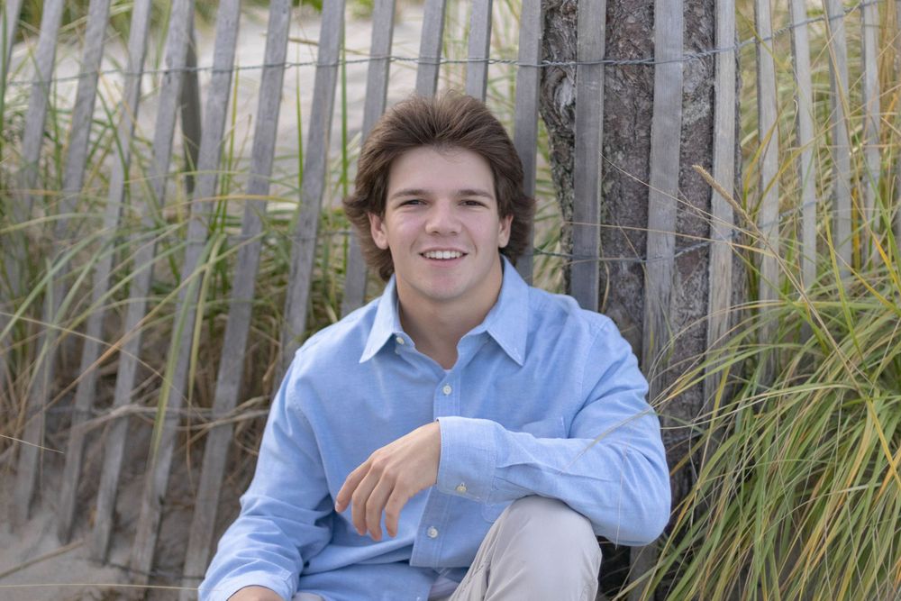 Smiling young man with wavy hair sitting against a wooden fence on a sandy beach.