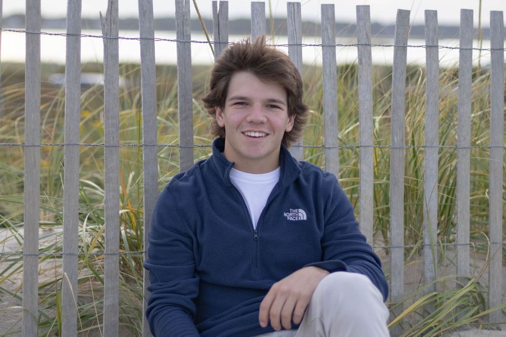 Young man in North Face jacket smiling outdoors by wooden fence with beach grass.