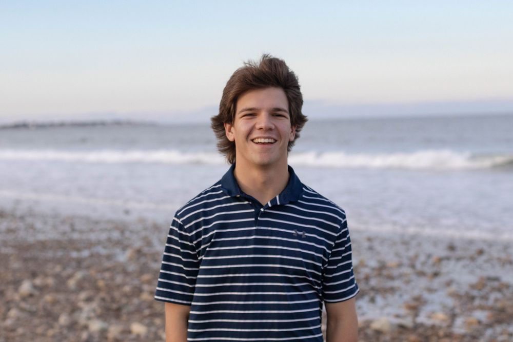 Smiling young man in striped polo shirt standing on a rocky beach at sunset.